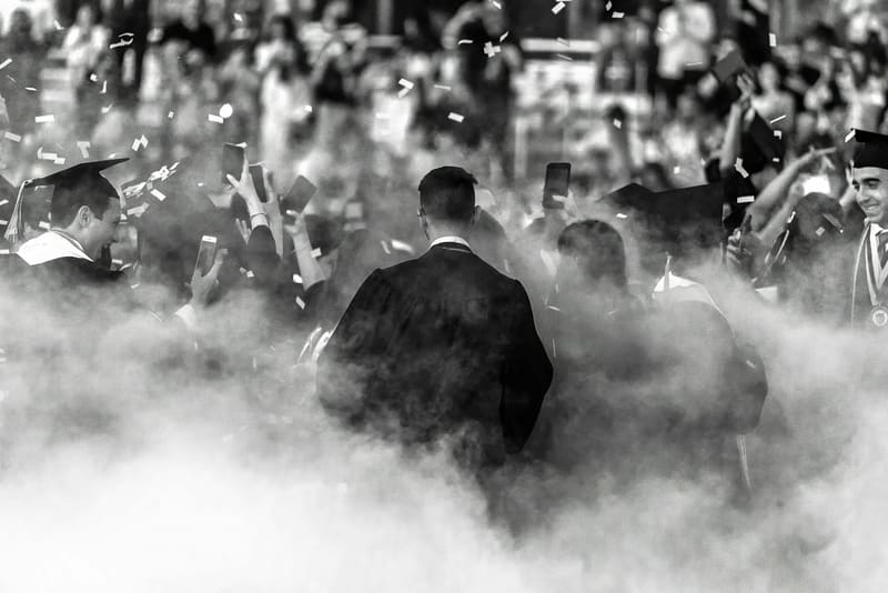 Graduates in gowns walking through confetti and smoke at a ceremony